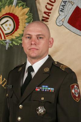 Bildē redzams Latvijas Nacionālās aizsardzības akadēmijas virsseržants, štāba virsseržants Jānis Timenieks svētku formā uz Mācību vadības pavēlniecības un Latvijas Nacionālās aizsardzības akadēmijas karogu fona